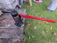 Photo showing the black heavy duty bottle jack with red handle attached, on a wooden surface outdoors next to grass.