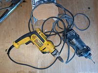Yellow DeWalt electric drill with cord and grinding bit, alongside a black rotary tool with a bit and attached cords on a wooden surface.