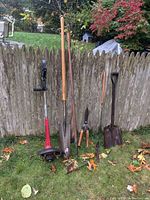 Full view of five garden tools leaning against wooden fence on grass with leaves, showing a red and black Toro trimmer edger, two shovels with wooden handles, a garden rake with metal tines and wood handle, and a weed scythe with wooden handle.