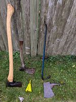 Lot of five garden and hand tools: two axes with wooden handles, an adze, a blue pry bar, and a yellow wedge resting on grass and against a wooden fence.