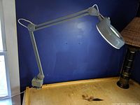 Gray industrial lamp with magnifier head mounted on adjustable arm, clamped to a wooden desk.
