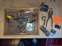 Wide shot of drawer and table with assorted machinist tools in a tray and on the surface, showing a variety of metal measuring tools and adjustable pliers.