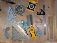 Full overview of the flat layout of measuring and layout tools on a wooden surface with tape measure, stud sensor, triangles, calk line, folding ruler, protractor, safety glasses, and plumb bob.