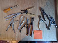 Photo shows a variety of pliers and a wooden handle screwdriver arranged on a wooden surface with an orange sticky note labeled '21P'.