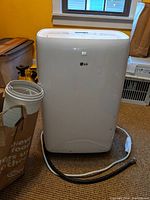 Front view of the portable LG air conditioner with the two hoses and power cord visible on the carpet next to it.