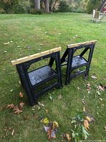 Two black plastic saw horses with yellow wooden tops on grass.