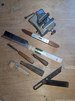 Photo showing small vise, Japanese saw, spirit level, chisels, angle squares, and marking tool on wooden surface.