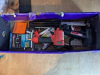 Box containing an electric stapler, hand stapler, air brad tool, and several boxes of staples and brads, photographed from above on wooden surface.