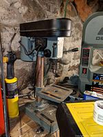 Mini drill press on a wooden surface in a basement setting, showing front and side views with the chuck and lever visible.
