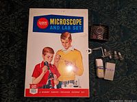 Original metal box with colorful vintage illustration on lid depicting two boys using the microscope, accompanied by some kit components on top