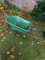 Wide angle view of green plastic wheelbarrow in yard showing overall condition and wooden handles.