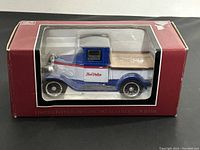 Front view of the die cast collector bank inside original maroon box with clear plastic window, showing the blue, white, and red 1928 Chevrolet National AB truck.
