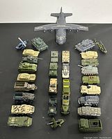 Top-down photo showing various die cast military vehicles and a large military transport airplane arranged in rows on a black surface.