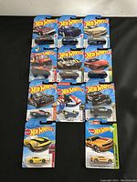 Overall photo of 11 die cast Hot Wheels cars in original packaging, showing front of each carded package arranged on black cloth.