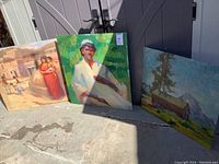 Three paintings leaning against an outdoor shed. From left to right: figurative painting with woman and child, portrait of man, landscape of cabin and mountains.