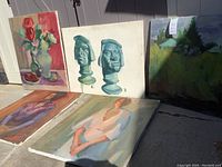 Five assorted art paintings on canvas leaning against a white shed wall, showing variety in subject and style.