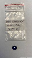 Plastic bag with handwritten label 'BLUE SAPPHIRE OVAL, FAR 8x10 K' and blue sapphire placed below the bag