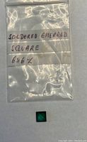 Photo shows a small square green emerald gemstone beneath a plastic bag labeled 'Soldered Emerald Square 6x6'.