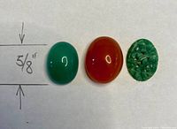 Three polished gemstones on white surface with handwritten scale showing 5/8 inch. Includes a green carved jade, green agate cabochon, and reddish agate cabochon.