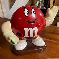 Photo of vintage M&M Red character candy dispenser, showing front with the smiling face, raised hand waving, and a candy held in the other hand, mounted on brown base.
