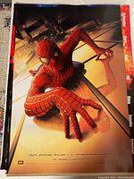 Photo of the front side of the original Spider-Man 2002 movie poster with Spider-Man crawling on glass of a skyscraper, bathed in warm light tones.