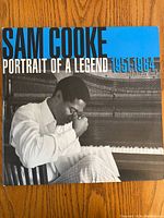 Front cover of Sam Cooke 'Portrait of a Legend 1951-1964' vinyl album showing an image of Sam Cooke sitting and smoking in front of a piano, with bold text.
