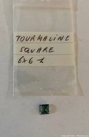 Photo showing a small square-shaped greenish-blue tourmaline gemstone next to a plastic bag labeled 'TOURMALINE SQUARE 6x6'.