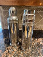 Two large vintage chrome thermoses placed side by side on a countertop with tiled backsplash.