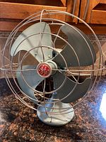 Front view of vintage GE metal desk fan with blue-gray blades and wire cage, showing the GE logo on the center hub.