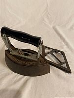 Side and front angled view of the vintage sad iron and accompanying metal trivet on a white cloth background.