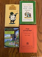 Four books arranged on wood surface: 'Toad of Toad Hall' with yellow illustrated cover, 'When We Were Very Young' hardcover with green slipcover, 'The Wind in the Willows' illustrated hardcover, and orange 'Toad of Toad Hall' play book.