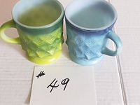 Two vintage Anchor Hocking Fire King Kimberley mugs placed side by side, showing front sides and handles, green/yellow and blue/white hues.