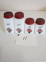 Front view of four Tiffin white milk glass spice shakers with red tops labeled Flour, Sugar, Salt, and Pepper with floral basket design.