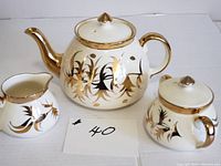Three piece tea set comprising teapot, creamer, and sugar bowl displaying gold and black decorative design on white ceramic.