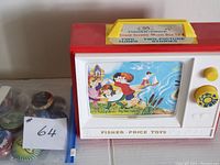 Photo of Fisher Price Giant Screen Music Box TV with red and white body, colorful illustrated screen, yellow knobs; next to a plastic bag containing 18 assorted Roxx toy pieces.