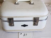 Front view of white textured hard travel/train case with metal clasps and handle, showing overall case shape and condition.