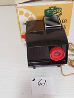 Photo showing the vintage brown View-Master projector with red lens, power cord visible, placed in front of original yellow and white box.