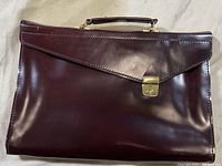 Front view of burgundy/maroon leather portfolio case with angled flap and gold clasp.