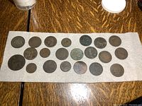 Full display of 21 antique foreign coins on a white cloth on wood surface, showing varied sizes and wear.