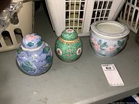 Three oriental themed ceramic items on a shelf: two ginger jars with lids (one green with Chinese motifs and one blue with floral vine design) and one small planter pot (blue with floral accents).