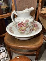 Porcelain pitcher with floral design seated inside matching wash basin, displayed on wooden chair