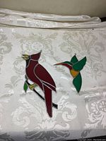 Two stained glass bird decorations on patterned fabric, showing vibrant red cardinal and green hummingbird.