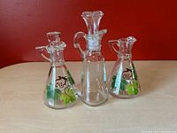 Three glass oil jars shown in front of a red background. Two conical jars with hand painted leaf motifs and the word Oil, one taller clear Anchor Hocking jar with stopper.