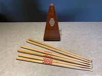 Wooden vintage Maelzel pyramid metronome with metal winder and dial plate with five pairs of wooden drumsticks placed in front on grey surface.