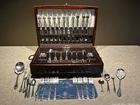 Open wooden case displaying flatware organized in fabric-lined slots and loose pieces on surface