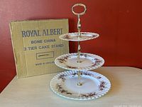 Three-tier cake stand shown assembled with original box behind it, clear floral pattern and gold accents visible
