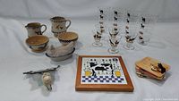 Lot overview showing pottery pitchers and bowls with animal motifs, blue spongware nesting hen, sets of vintage animal themed glassware, cow stained glass nightlight, Lillian Vernon cow tile trivet, and Le Chat Noir coasters.