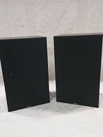 Pair of Boston Acoustics speakers shown side by side on a quilted white surface, both with black fabric grills and Boston Acoustics logo at the bottom.