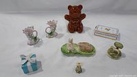 Seven decorative ceramic items arranged on a white fabric background including the brown bear baby bank, blue Tiffany gift box trinket, two pink flower candleholders, horse figurine, and bunny figurines.