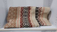 Blanket folded on a white shelf showing detailed geometric patterns in earth tones including red, black, and brown.